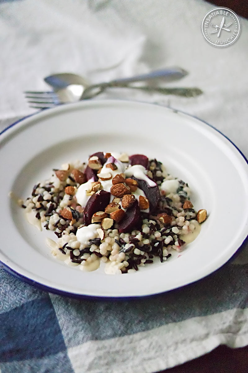 A salad of black rice and pearl cous cous, served with a roasted garlic and tahini dressing, greek yoghurt, baby beetroot and smoked almonds. Here, it is served on a blue-rimmed tin plate and laid next to a fork and spoon, on a blue tea towel.
