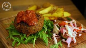 Lentil patty with a red sauce sits on fresh rocket leaves with shredded red cabbage and chips on the side.