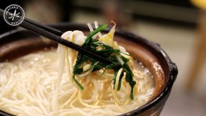 Tender white rice noodles sit in a rich creamy broth, mixed with some greens and bean sprouts. A chopstick gently lifts a mouthful out of the soup.
