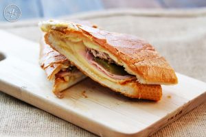 A cuban sandwich - mustard, swiss cheese, pickles ham, braised pork shoulder - is halved and sits on a wooden board.