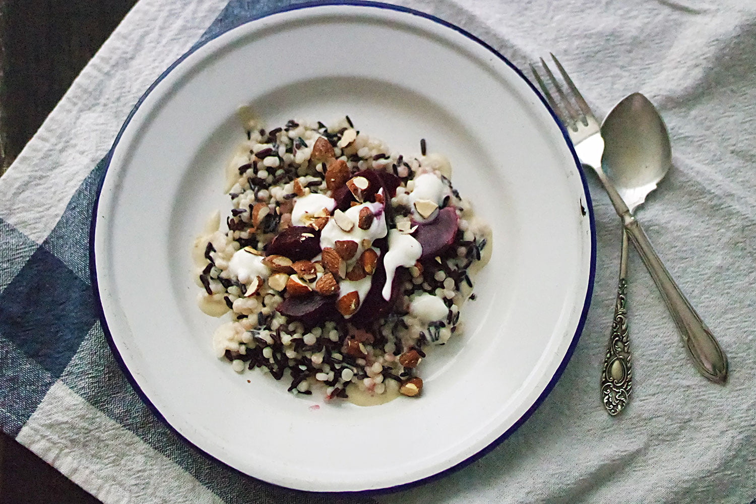 A salad of black rice and pearl cous cous, served with a roasted garlic and tahini dressing, greek yoghurt, baby beetroot and smoked almonds. Here, it's served on a blue-rimmed tin plate and laid next to a fork and spoon, on a blue tea towel.