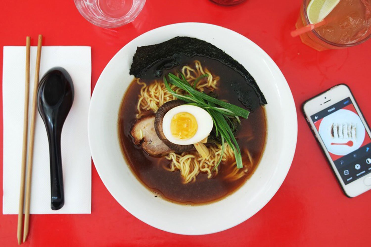 A bowl of "the darkness" ramen from Rising sun workshop, photographed aerially with cutlery and a mobile phone flanking it from either side.