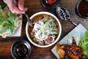 A fork digging into a bowl of pho amongst a table of food that includes grilled chicken