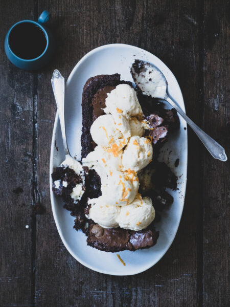 A loaf of chocolate brownie topped with a mound of ice cream, garnished with orange zest. Served with a side of coffee
