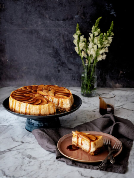 A caramel apple cheese cake on a cake stand, with a slice served on the side.