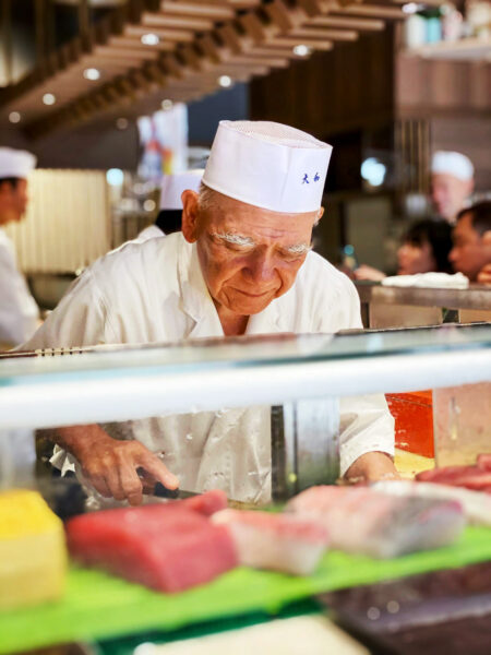 An elderly sushi chef behind the counter, preparing sushi
